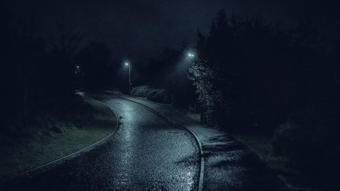 Une route de campagne mouillée qui tourne dans la nuit, éclairée par deux lampadaires isolés.
