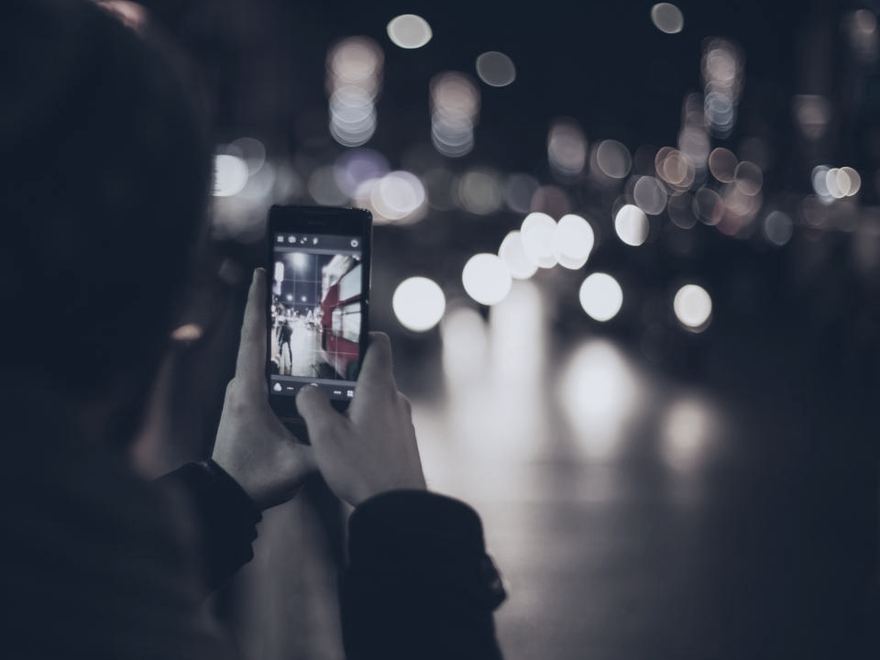 Deux mains tiennent un téléphone allumé dans une rue de nuit, les lumières floues en arrière-plan.